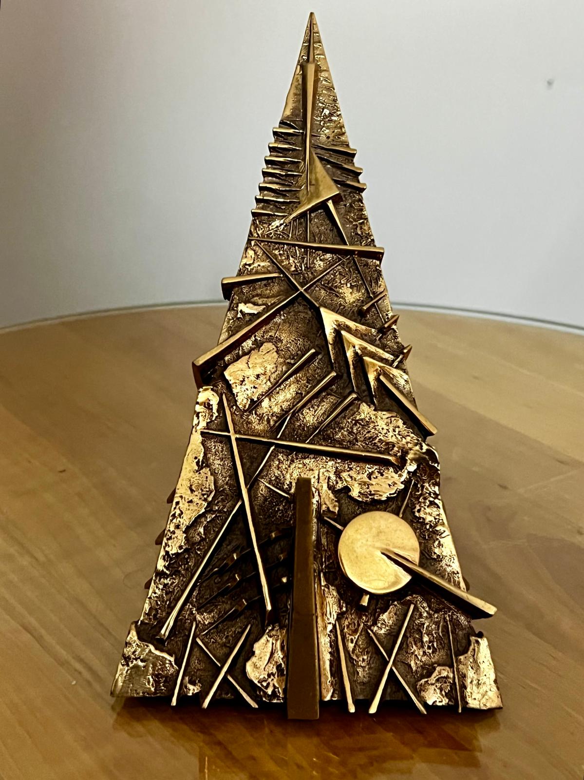 La scultura intera vista di fronte: una guglia di bronzo dorato dalla superficie lacerata, con un disco in basso a destra.